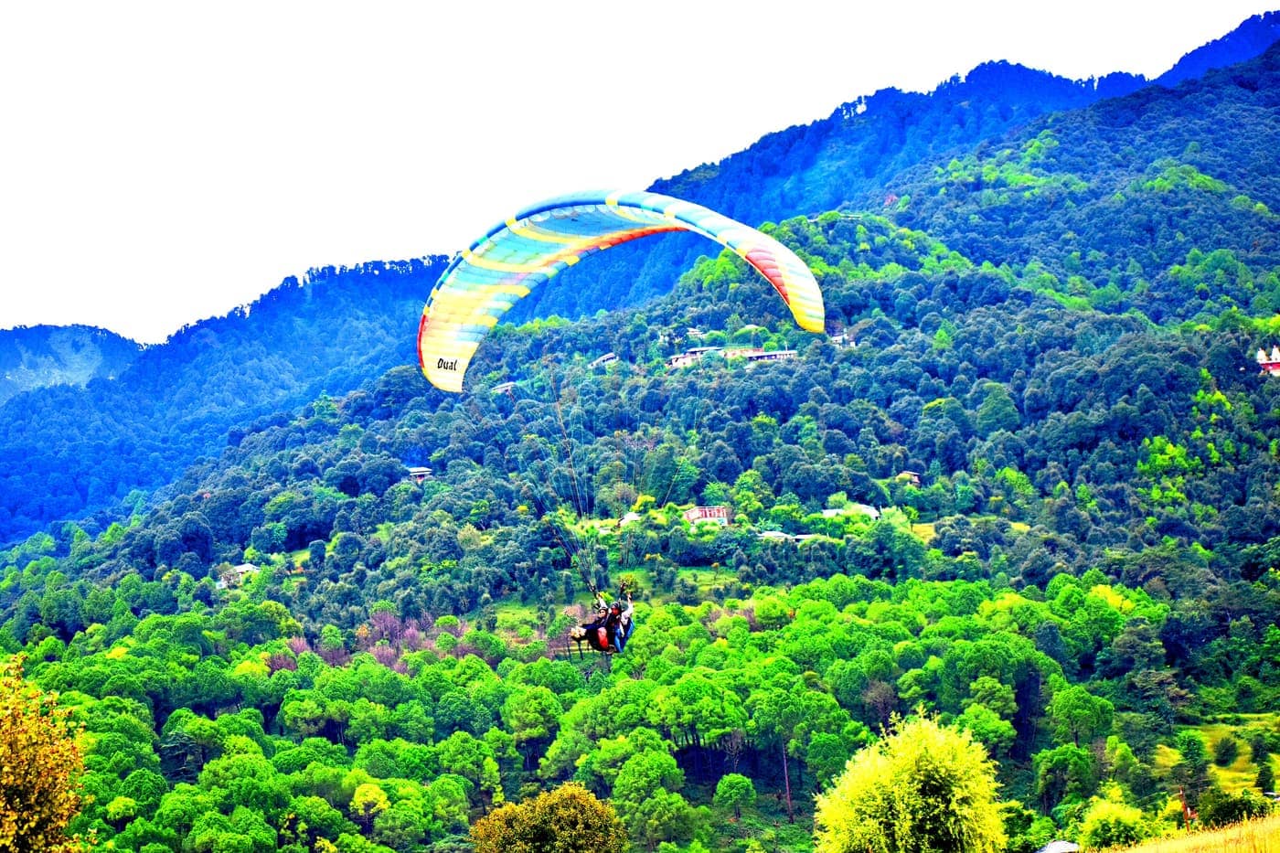 A tandem paraglider flying over green forested hills at Bir Billing